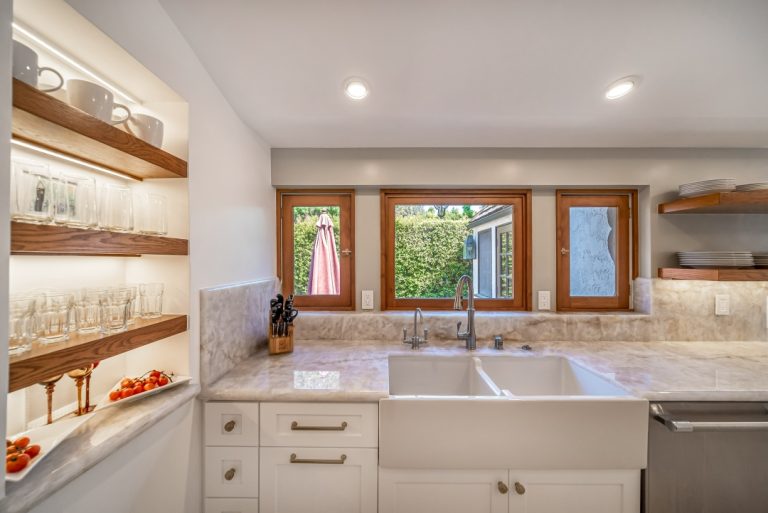Farmhouse sink beneath wood-framed windows beside open display shelves.