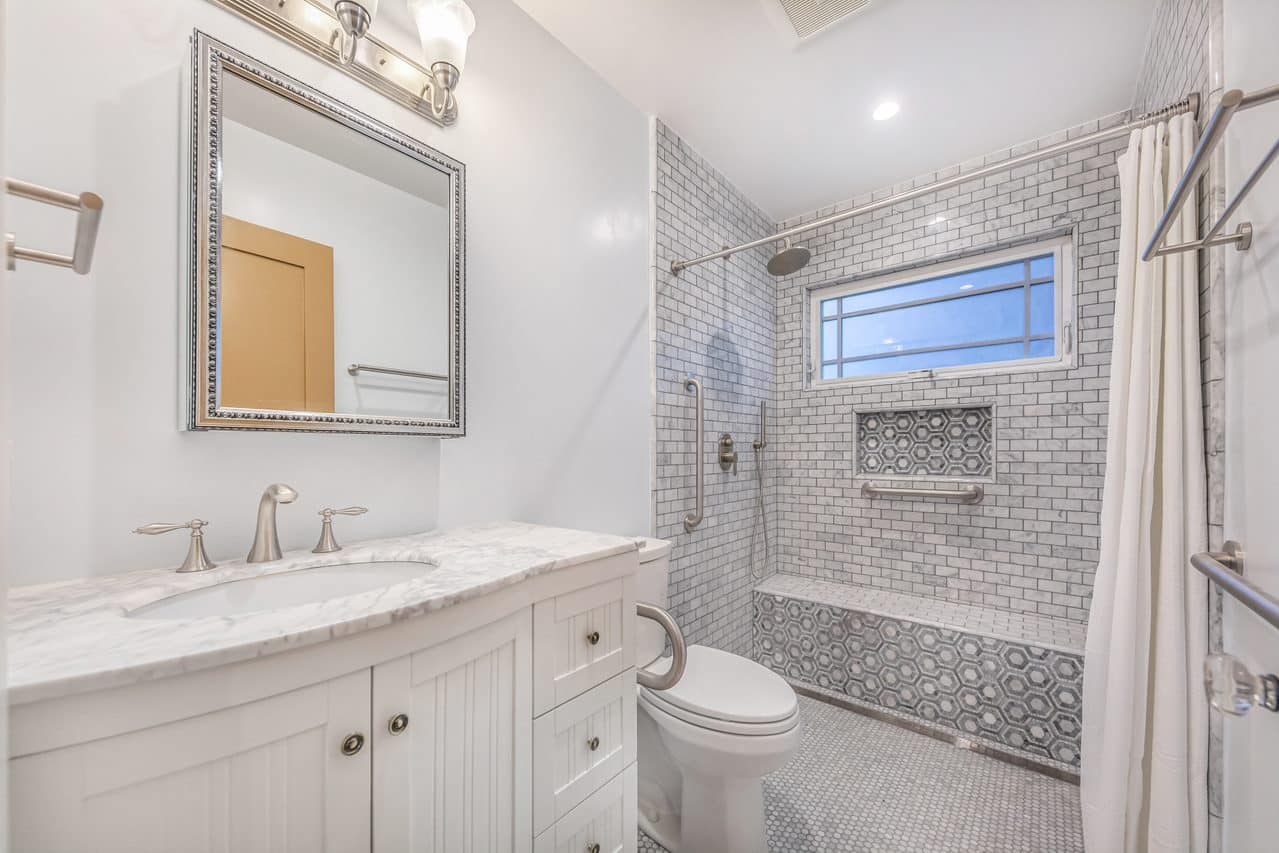 White bathroom vanity beside a shower with a window and bench.